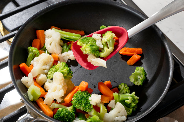 Vegetables including broccoli, cauliflower, and carrots being sautéed in a pan with a Cuisipro red Silicone Spoon with a stainless steel handle