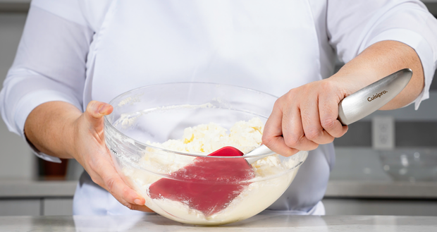 Person using a red Cuisipro spatula to blend ingredients in a glass bowl.